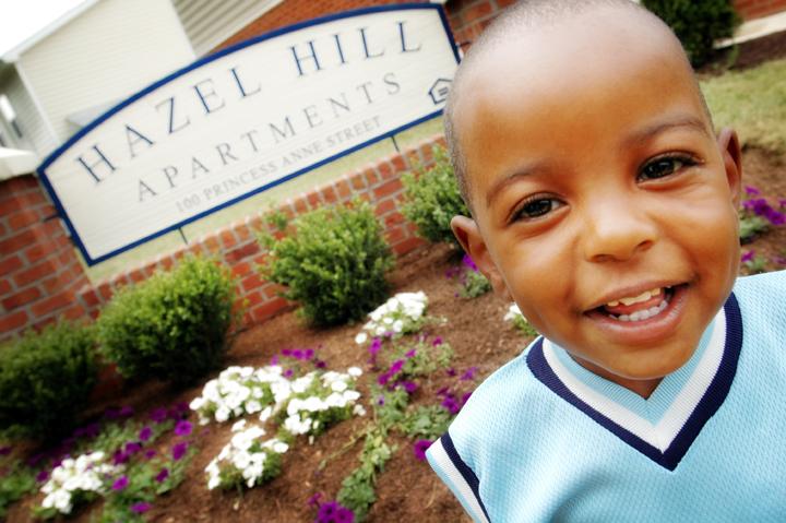 young boy smiling at the camera wearing a light blue vest in front of a sign for Hazel Hill apartments